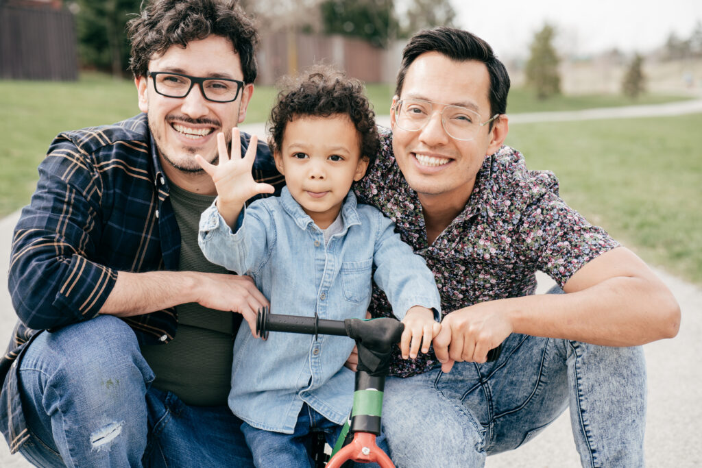two fathers with their young son between them on a scooter, smiling at camera