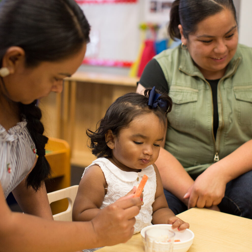 Two teachers sitting at table with toddler in classroom setting